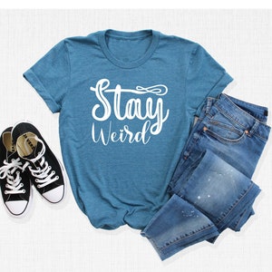 以下が含まれることがあります： 白い文字で「Stay Weird」と書かれたターコイズブルーのTシャツ。Tシャツは白い表面に置かれ、黒いスニーカーと青いジーンズが置かれている。