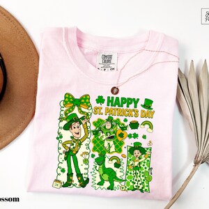 A pink t-shirt with a cartoon design of characters from Toy Story, including Woody and St. Patrick's Day-themed elements, is displayed alongside a hat and a fan.