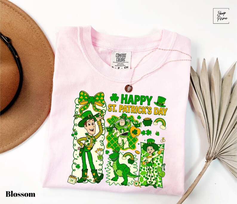 A pink t-shirt with a cartoon design of characters from Toy Story, including Woody and St. Patrick's Day-themed elements, is displayed alongside a hat and a fan.