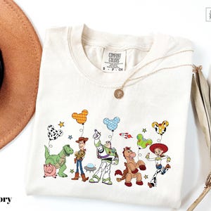 A white t-shirt with a cartoon illustration of various characters from the Toy Story franchise, including Woody, Buzz Lightyear, and other beloved characters.
