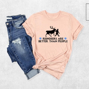 May include: A pink t-shirt with a black silhouette of a reindeer and a person walking. The text on the shirt reads "Reindeers are better than people" with a blue snowflake on each side of the text.