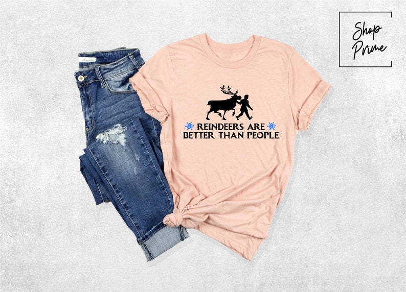 May include: A pink t-shirt with a black silhouette of a reindeer and a person walking. The text on the shirt reads "Reindeers are better than people" with a blue snowflake on each side of the text.