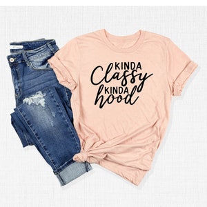 May include: A light pink t-shirt with the text "Kinda Classy Kinda Hood" in black lettering. The shirt is shown on a white background with a pair of blue jeans.