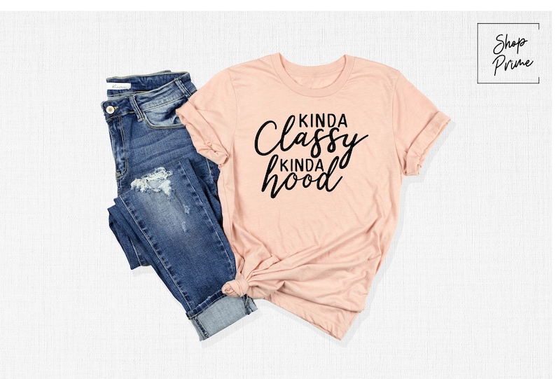 May include: A light pink t-shirt with the text "Kinda Classy Kinda Hood" in black lettering. The shirt is shown on a white background with a pair of blue jeans.