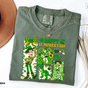 A green t-shirt with a cartoon character and text printed on it, along with a hat and some leaves, all placed on a white surface.