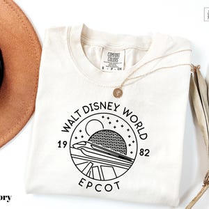 May include: Ivory-colored t-shirt with a black graphic of the Walt Disney World EPCOT logo. The logo includes the text "WALT DISNEY WORLD", "EPCOT", and the numbers "1982". A gold necklace and a tan hat are also visible.