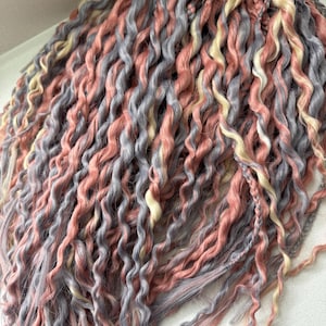May include: A bundle of synthetic hair extensions in shades of pink, gray, and blonde. The hair is in a wavy, curly style.