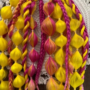 Könnte beinhalten: Nahaufnahme von farbenfrohem Haarschmuck. Geflochtene und mit Perlen besetzte Strähnen in leuchtenden Gelb-, Rosa- und Orangetönen. Die Accessoires sind in einem vertikalen Muster angeordnet, das die komplizierten Details des handgefertigten Designs zeigt. Die leuchtenden Farben erzeugen einen verspielten und auffälligen Look.