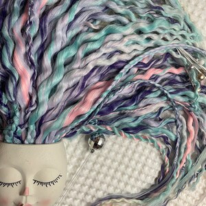 May include: A close-up of a bundle of colorful synthetic hair extensions in shades of pink, blue, purple, and white. The hair is arranged in a loose, flowing style, with some strands cascading down.