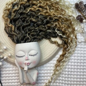 May include: A white ceramic planter shaped like a girl's face with closed eyes and pink lips. The planter has long, curly brown and blonde hair.