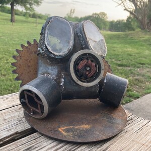 May include: A rusty metal gas mask sculpture with a circular base and a gear on the side. The mask has two round eye pieces and a circular filter. The sculpture is on a wooden surface.