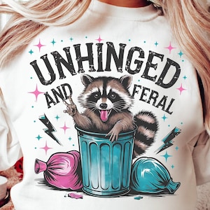 Pode incluir: Camisola branca com um gráfico de um guaxinim num caixote azul. O texto "Unhinged and Feral" está impresso na camisola.