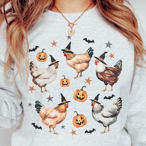 May include: A light gray sweatshirt featuring a Halloween-themed design. The design includes illustrations of chickens wearing witch hats, orange jack-o'-lanterns, bats, and stars. The chickens are in various colors, and the overall style is whimsical.