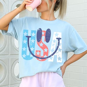 Op de afbeelding: Een lichtblauw T-shirt met een smiley-ontwerp dat het woord "USA" in rood, wit en blauw bevat. Het wordt gecombineerd met lichtpaarse shorts. De afbeelding toont een casual zomeroutfit.