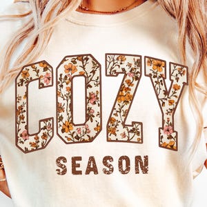 Puede incluir: Camiseta color crema con las palabras "COZY SEASON" en una fuente marrón llena de flores. Las letras están delineadas en marrón y las flores son en tonos rosa, naranja y amarillo. La camiseta tiene un aspecto vintage, ligeramente descolorido.