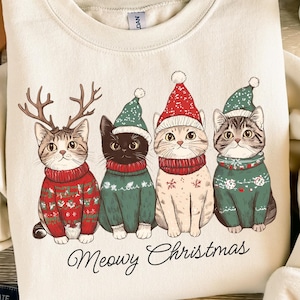 May include: A cream-colored sweatshirt with a graphic of four cats wearing Christmas hats and sweaters. The cats are wearing reindeer antlers, a Santa hat, a green hat, and a red hat. The text "Meowy Christmas" is printed below the cats.