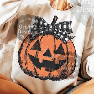 May include: Cream-colored sweatshirt featuring a distressed orange jack-o'-lantern graphic with a black and white plaid bow. The pumpkin has a carved face with triangular eyes and a smiling mouth. The words "Original Designer" are printed on the sweatshirt.