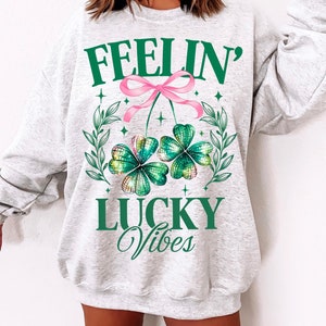 May include: A gray sweatshirt with the text "Feelin' Lucky Vibes" and a green four-leaf clover with a pink bow.