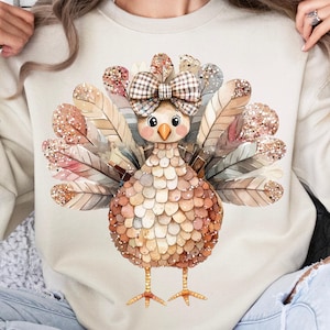 May include: A beige sweatshirt with a watercolor illustration of a turkey with a plaid bow on its head. The turkey has brown and gold feathers and a glittery texture.