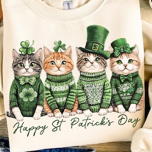 May include: A beige sweatshirt with a graphic of four cats wearing green sweaters and hats. The cats are lined up and the text "Happy St. Patrick's Day" is printed below them.