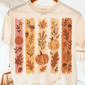 Puede incluir: Top corto color crema con un diseño de temática otoñal. El diseño incluye rayas verticales con calabazas, hojas y bellotas en tonos naranja, marrón y rosa. La camiseta está colgada de una percha de madera.
