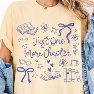 以下が含まれることがあります： 「Just One More Chapter」の青い文字がプリントされた、薄い黄色のTシャツ。本、花、ハート、リボン、メガネ、コーヒーカップの青いイラストが描かれています。袖は折り返されています。