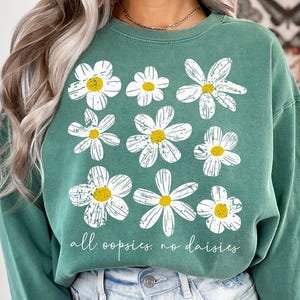 May include: Green sweatshirt featuring a daisy floral pattern with yellow centers and white petals. The text "all oopsies, no daisies" is printed in a cursive font below the flowers. The sweatshirt is made of a soft material.