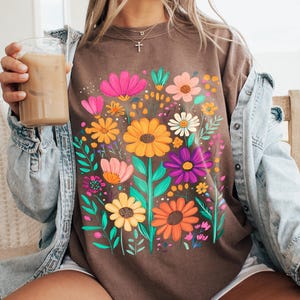 May include: Brown t-shirt with a colourful floral design, including orange, yellow, pink, and purple flowers. The graphic tee has green leaves and stems. The model is wearing a denim jacket and white shorts.