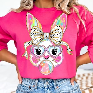May include: A pink sweatshirt with a colorful cartoon bunny wearing heart-shaped glasses and a bow. The bunny is holding a colorful Easter egg.