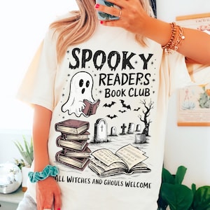以下が含まれることがあります： 白いTシャツに、本を読んでいる幽霊、本の積み重ね、墓地、コウモリが描かれた黒いグラフィックデザインが施されています。シャツには「Spooky Readers Book Club」と「All Witches and Ghouls Welcome」の文字が書かれています。