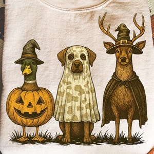 May include: Cream-colored sweatshirt featuring a Halloween-themed graphic. The graphic depicts a duck in a witch hat and pumpkin, a dog in a ghost costume, and a deer in a witch hat and cape. The illustration is in a vintage style.