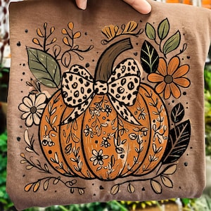 May include: Brown t-shirt with an orange pumpkin illustration. The pumpkin has a leopard print bow and floral details, surrounded by leaves and flowers. A seasonal design, ideal for autumn.