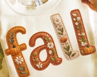 May include: Cream-coloured sweatshirt featuring the word "fall" embroidered in brown, beige, and orange. The letters are decorated with pumpkins, flowers, and leaves, creating an autumnal theme.