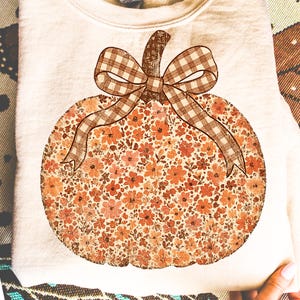 May include: Cream-colored sweatshirt featuring a large pumpkin design. The pumpkin is filled with a floral pattern in shades of orange and peach, with a brown gingham bow and stem. A cozy fall-themed apparel item.