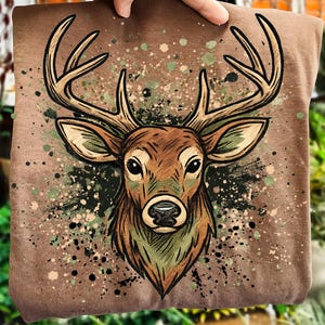 以下が含まれることがあります： 茶色のTシャツ。鹿の頭と角の精巧なイラストが描かれています。鹿は茶色、ベージュ、緑色で描かれ、迷彩柄の背景が施されています。デザインはTシャツの中央に配置されています。