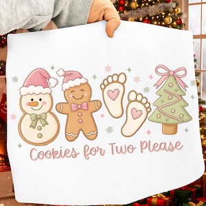 Puede incluir: Funda de almohada blanca con temática navideña con un muñeco de nieve, un hombre de jengibre, huellas de bebé con corazones y un árbol de Navidad. El texto "Cookies for Two Please" está en rosa. Festivo y de temporada.