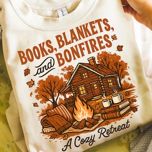 May include: Cream-coloured sweatshirt with a graphic featuring a cabin, bonfire, books, and blankets. The text reads "BOOKS, BLANKETS, and BONFIRES" and "A Cozy Retreat" in brown. Autumn leaves and mugs of coffee complete the design.
