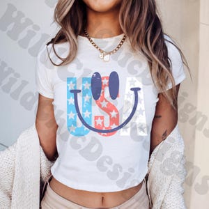 Op de afbeelding: Wit T-shirt met een patriottisch smiley-gezicht ontwerp. Het ontwerp bevat de letters "USA" met blauwe en rode sterren en strepen, wat een vrolijke en feestelijke uitstraling cre&euml;ert. Het shirt is een klassieke ronde hals.