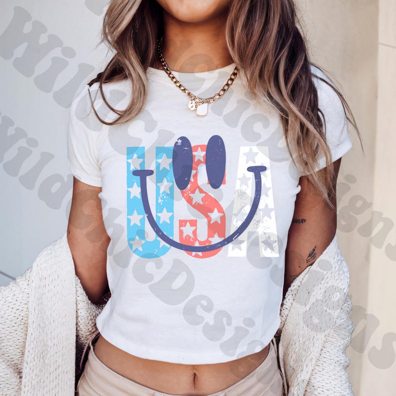 Op de afbeelding: Wit T-shirt met een patriottisch smiley-gezicht ontwerp. Het ontwerp bevat de letters "USA" met blauwe en rode sterren en strepen, wat een vrolijke en feestelijke uitstraling cre&euml;ert. Het shirt is een klassieke ronde hals.