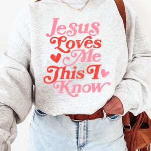 Puede incluir: Una sudadera gris con el texto "Jesus Loves Me This I Know" en letras rosa y rojas con un gr&aacute;fico de coraz&oacute;n.