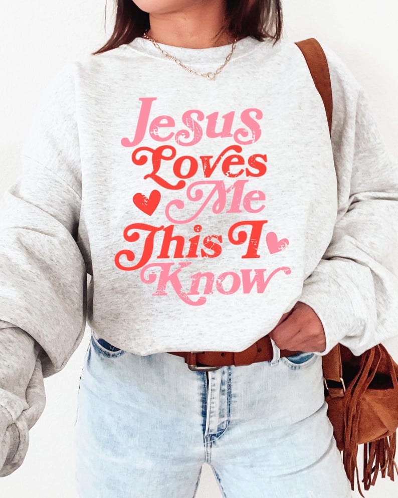 Puede incluir: Una sudadera gris con el texto "Jesus Loves Me This I Know" en letras rosa y rojas con un gr&aacute;fico de coraz&oacute;n.