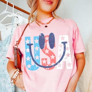 Op de afbeelding: Een lichtroze t-shirt met het woord "USA" in een smiley-ontwerp. De letters zijn in rood, wit en blauw met steraccenten. Het shirt heeft korte mouwen.