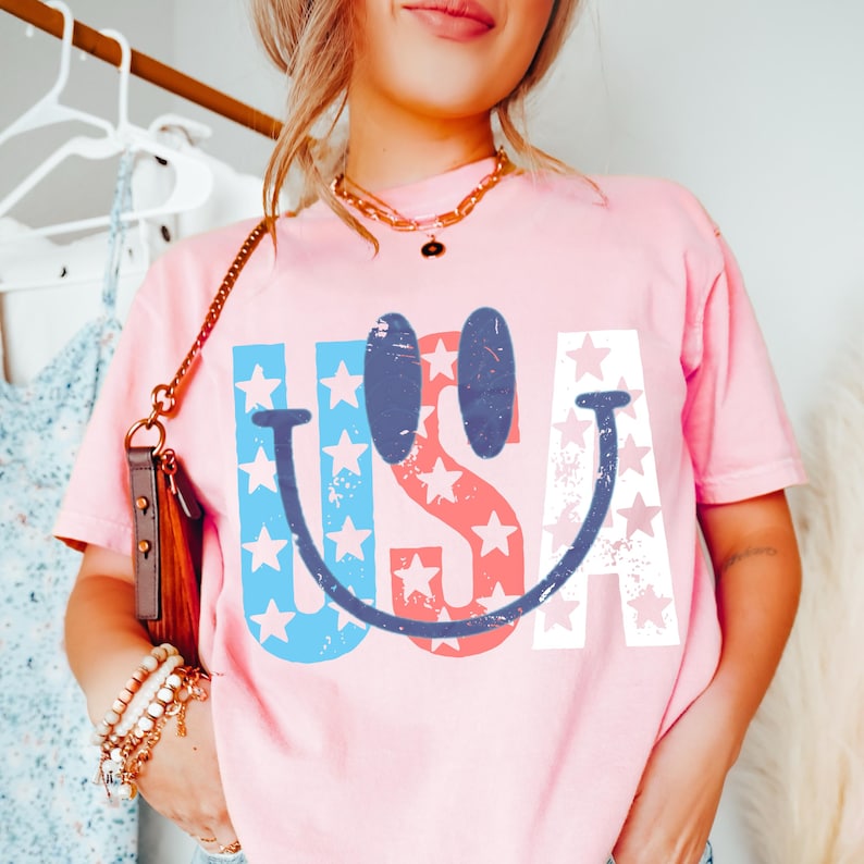 Op de afbeelding: Een lichtroze t-shirt met het woord "USA" in een smiley-ontwerp. De letters zijn in rood, wit en blauw met steraccenten. Het shirt heeft korte mouwen.
