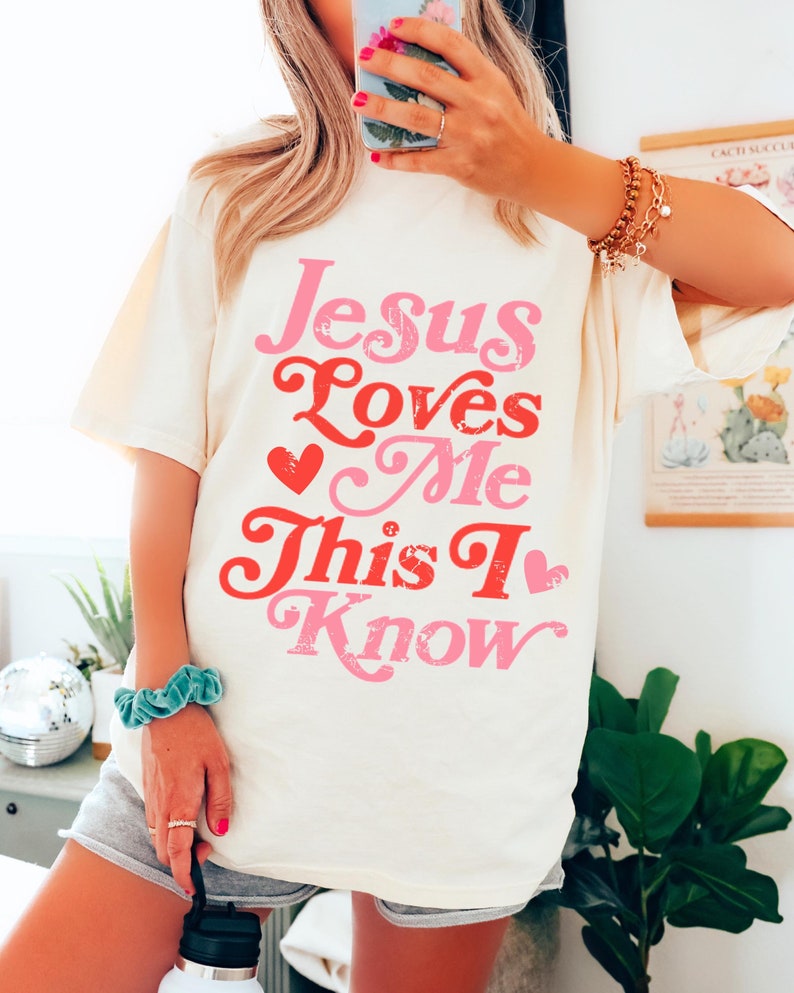 Puede incluir: Una camiseta blanca con un gr&aacute;fico rosa y rojo que dice "Jesus Loves Me This I Know" con corazones.