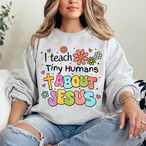 May include: A gray sweatshirt with a colorful graphic that says "I teach tiny humans about Jesus" with flowers and hearts around the text.