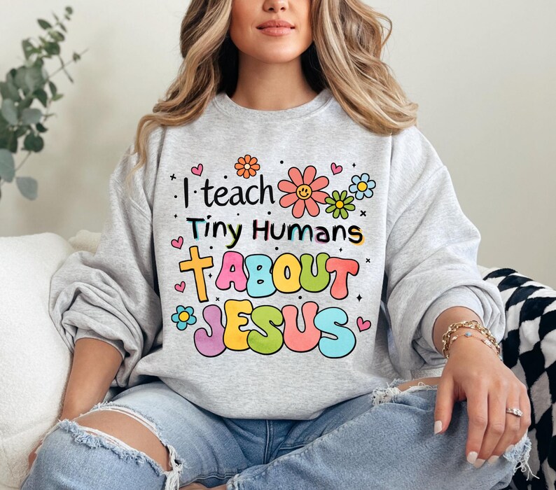 May include: A gray sweatshirt with a colorful graphic that says "I teach tiny humans about Jesus" with flowers and hearts around the text.