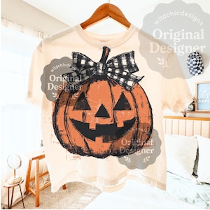 May include: Cream-colored t-shirt featuring a distressed orange jack-o'-lantern graphic with a black and white plaid bow. The design includes a smiling face and is perfect for Halloween. The text "Original Designer" is also visible.