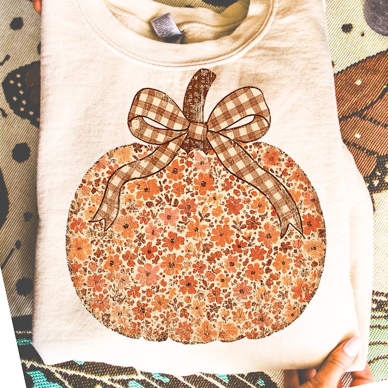 May include: Cream-colored sweatshirt featuring a large pumpkin design. The pumpkin is filled with a floral pattern in shades of orange and peach, with a brown gingham bow and stem. A cozy fall-themed apparel item.