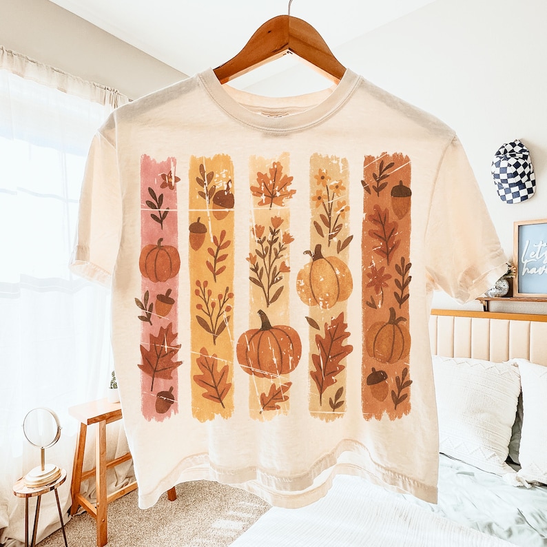 May include: Cream-colored crop top featuring a fall-themed design. The design includes vertical stripes with pumpkins, leaves, and acorns in shades of orange, brown, and pink. The shirt is hanging on a wooden hanger.