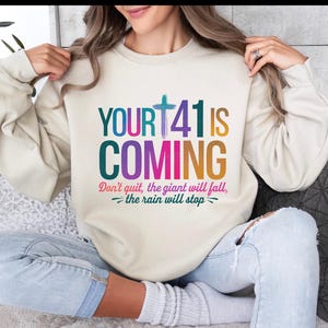 May include: A cream colored sweatshirt with the text "YOUR+41 IS COMING" in rainbow colors. The text "Don't quit, the giant will fall, -the rain will stop- " is printed below the main text.